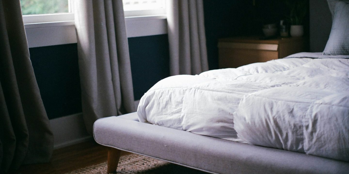 Cama de casal com edredão branco junto a uma janela, num quarto com cortinas cinzentas