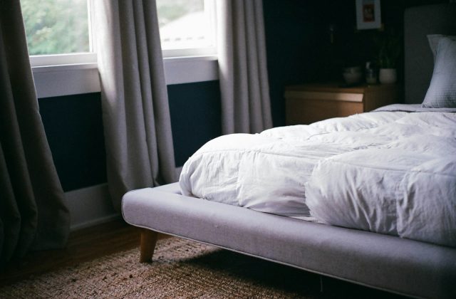 Cama de casal com edredão branco junto a uma janela, num quarto com cortinas cinzentas