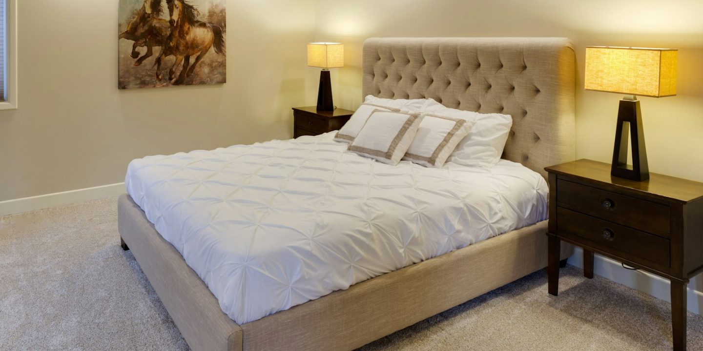 Cama de casal com cabeceira estofada bege, edredão branco e espaço livre dos dois lados, entre duas mesas de cabeceira