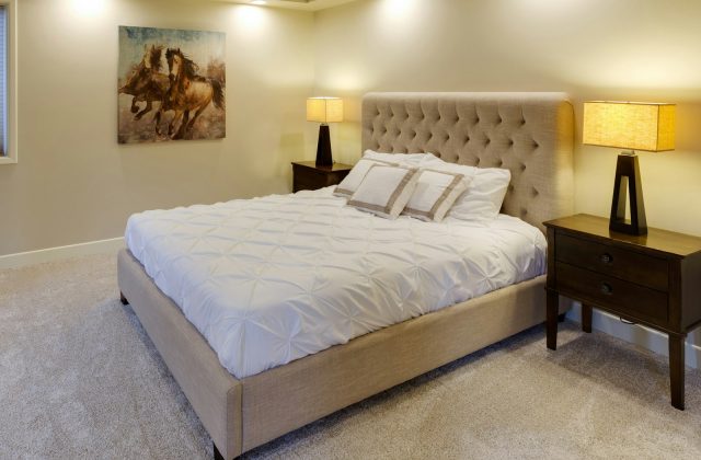 Cama de casal com cabeceira estofada bege, edredão branco e espaço livre dos dois lados, entre duas mesas de cabeceira